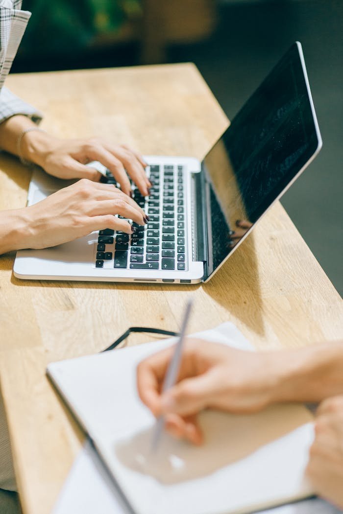 Hands typing on a laptop and writing in a notebook at a desk, emphasizing teamwork.