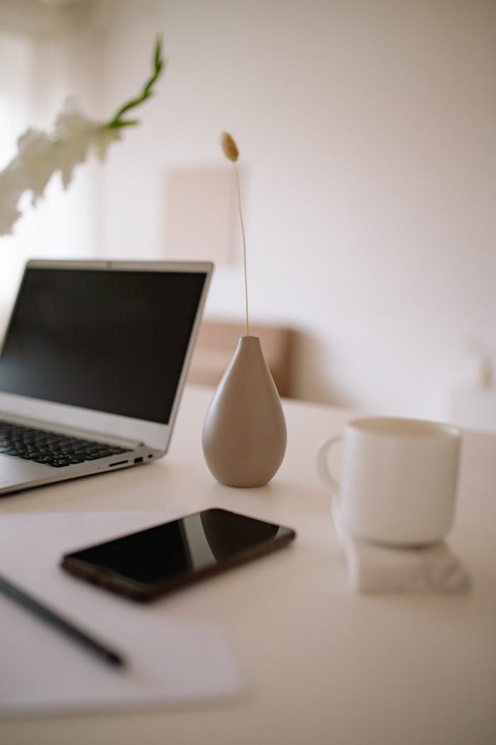 A clean and modern workspace featuring a laptop, vase, cup, smartphone, and paper on a white table.